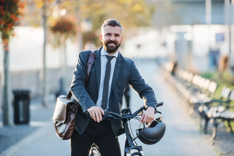 Ein lächelnder Mann in Businesskleidung, mit Sakko und Krawatte, steht draußen neben einem Fahrrad. Er trägt eine Umhängetasche und hält einen Fahrradhelm in der Hand, während die herbstliche Umgebung und eine Allee mit Bänken im Hintergrund sichtbar sind.