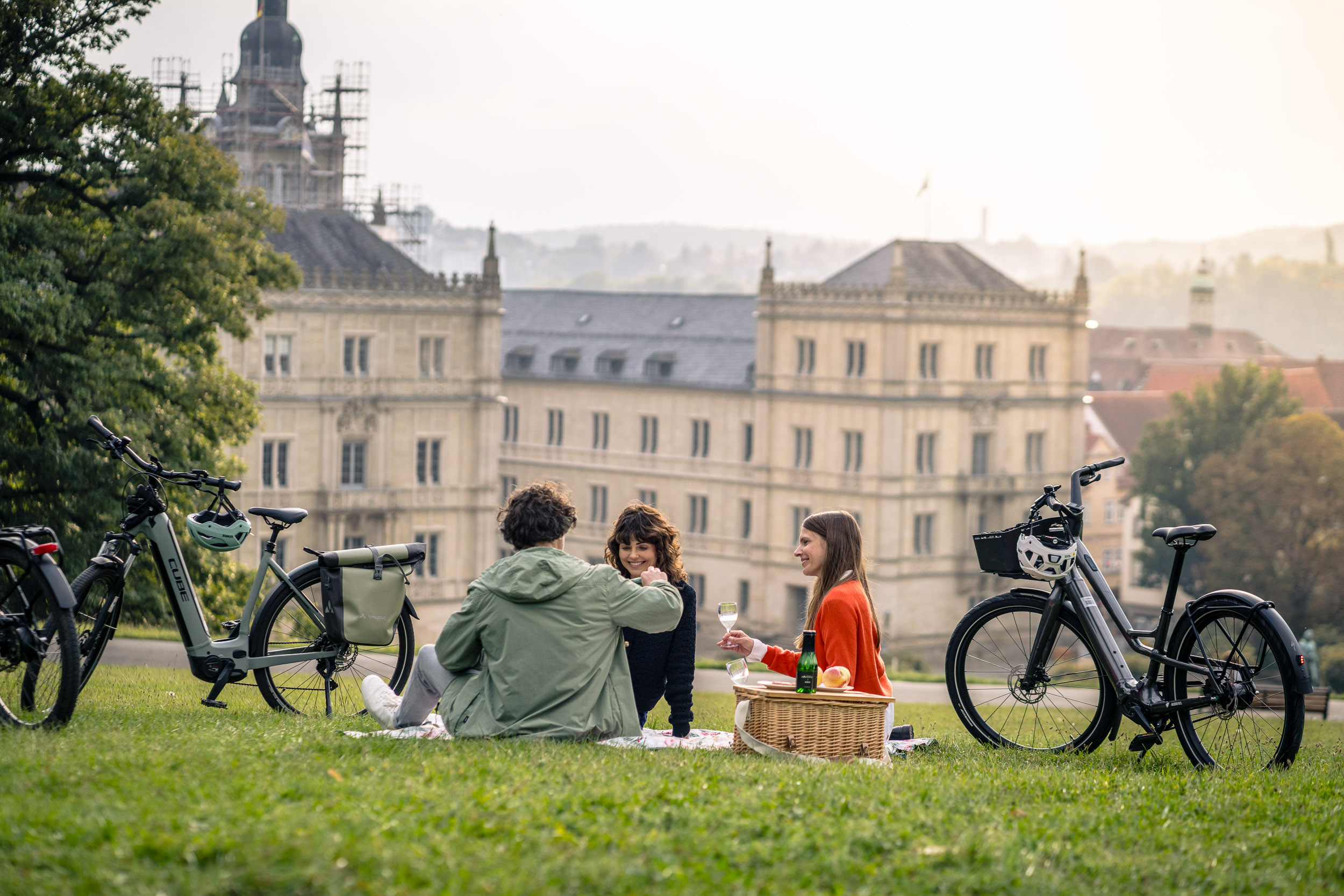Drei Personen machen ein Picknick auf einer Wiese mit E-Bikes im Vordergrund und einem historischen Gebäude im Hintergrund.