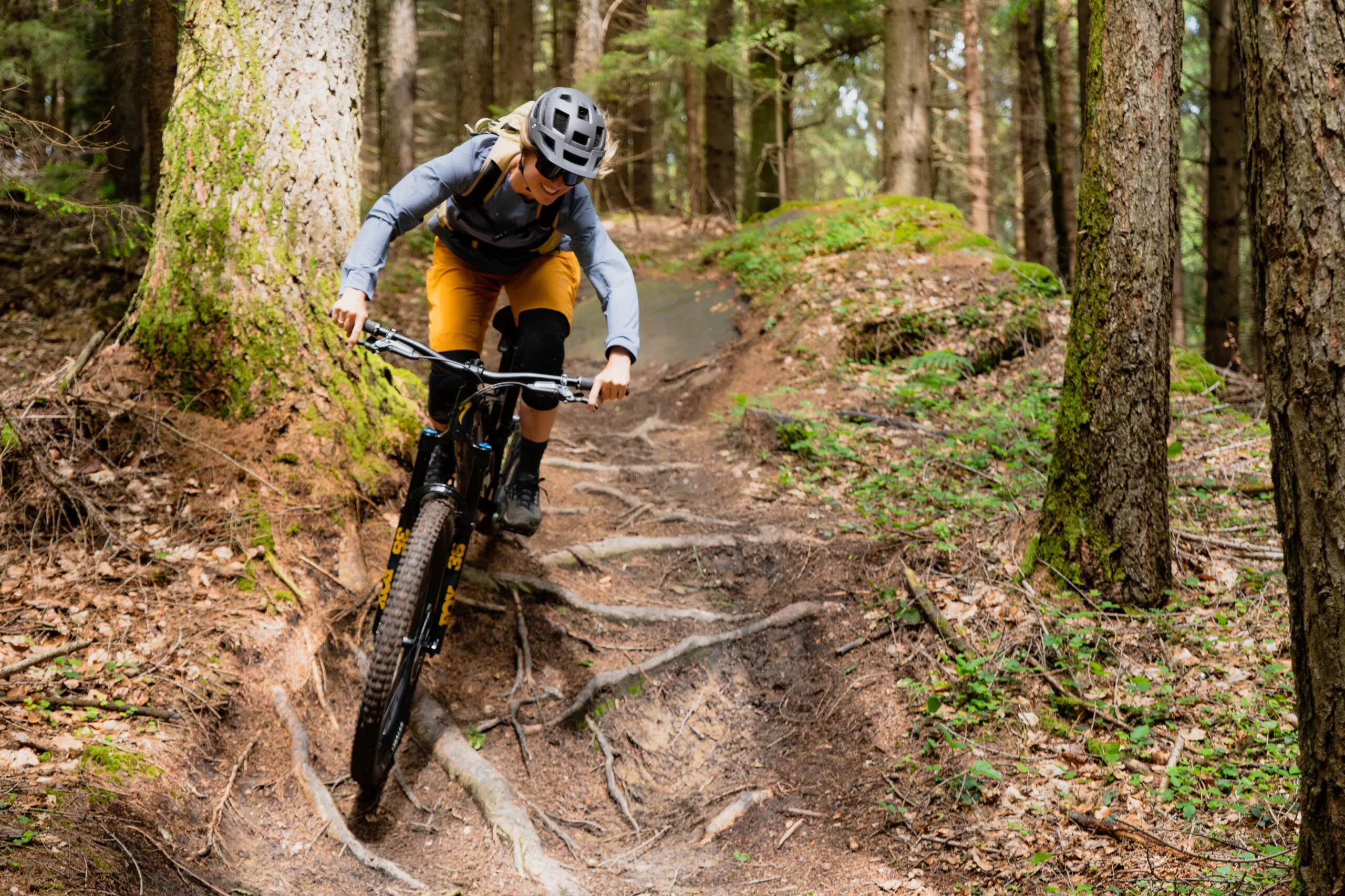 Eine Mountainbikerin fährt einen steilen, verwurzelten Waldpfad hinunter. Die Fahrerin trägt Helm, Rucksack und gelbe Shorts, während sie sich konzentriert durch die kurvige und anspruchsvolle Strecke bewegt.