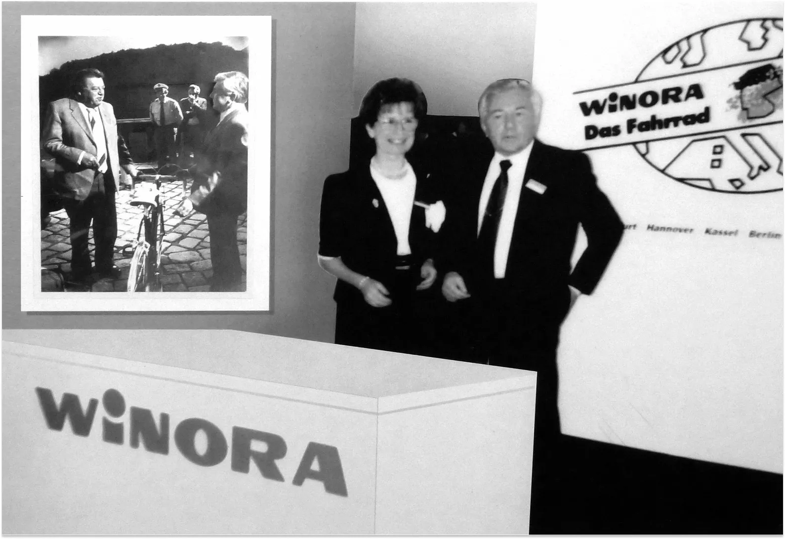 Schwarz-Weiß-Foto von zwei Personen in formeller Kleidung vor einem Messestand mit dem Logo 'Winora – Das Fahrrad' und einem gerahmten Bild im Hintergrund.