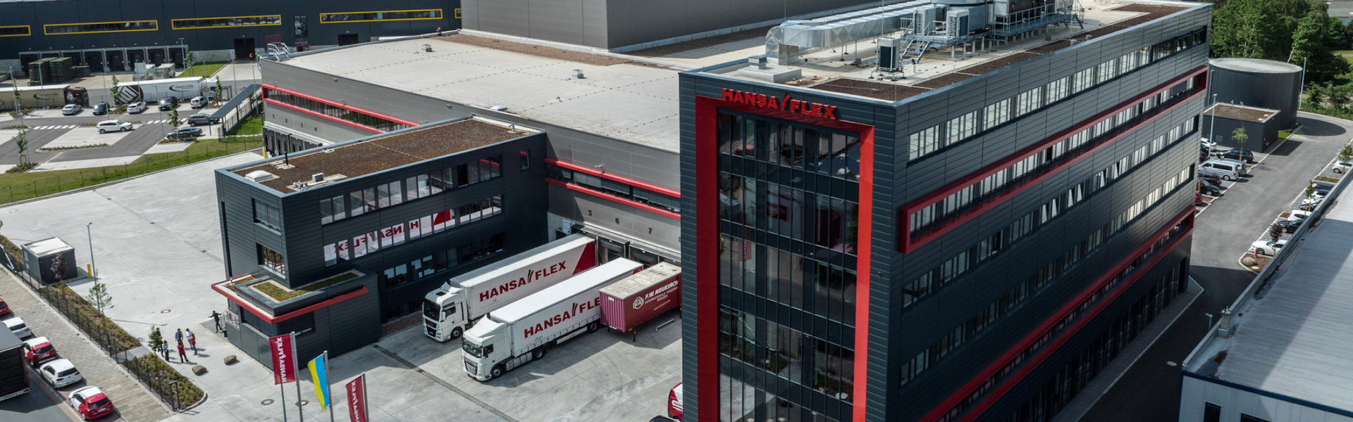 Aerial view of a modern industrial building with the words "Hansa Flex" on the facade. Two white trucks with the same logo are parked at loading ramps. The site is spacious and surrounded by parked cars and other industrial facilities.