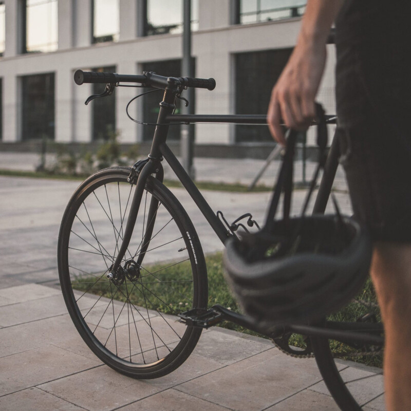 A person is holding a bicycle helmet in their hand and standing next to a black bike on a paved path.