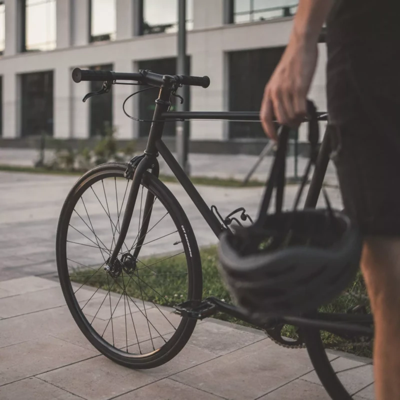 Eine Person hält einen Fahrradhelm in der Hand, während sie neben einem schwarzen Fahrrad steht. Im Hintergrund ist ein modernes Gebäude mit großen Fenstern zu sehen.