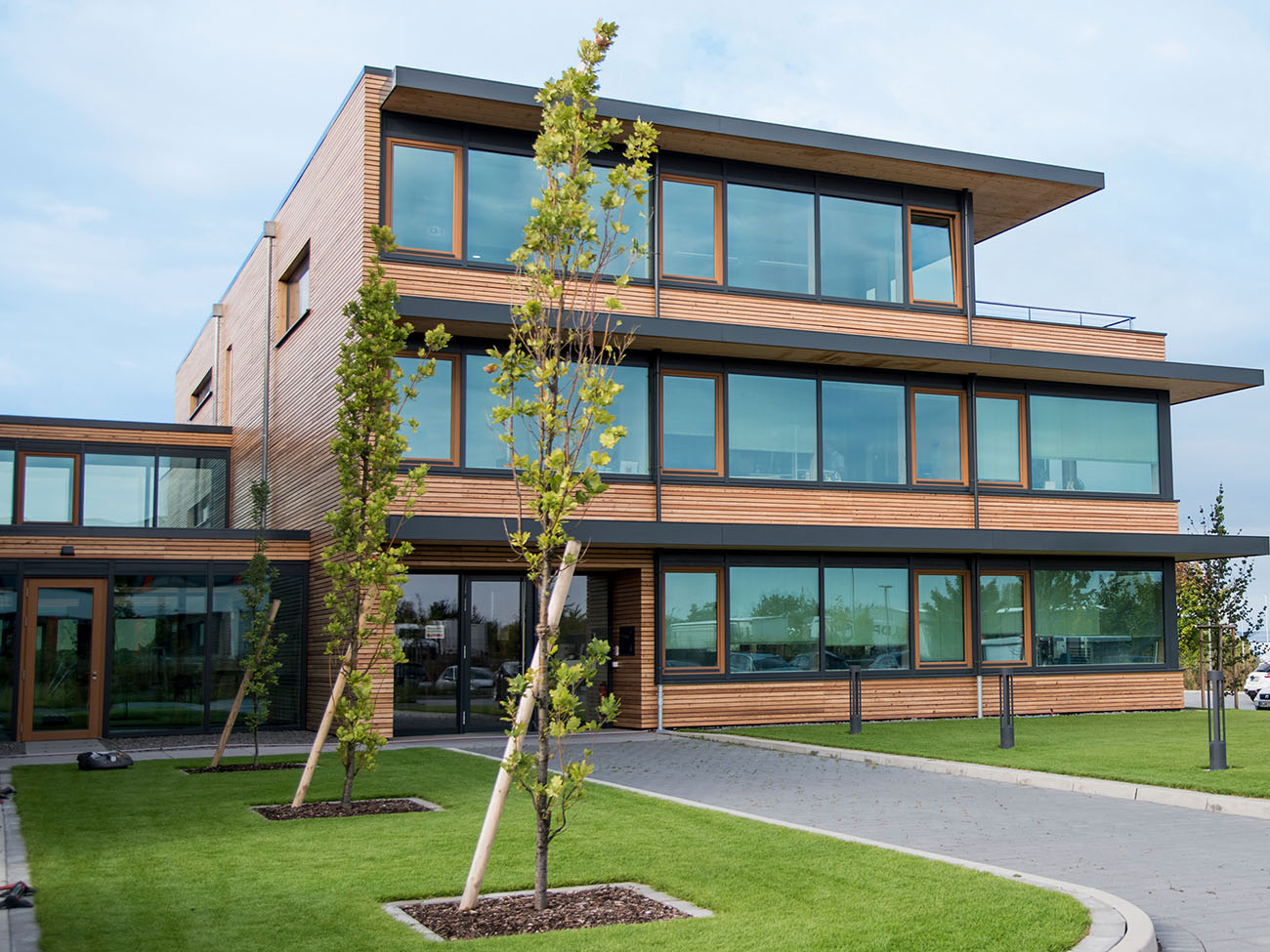 A modern, three-story building with large glass windows and a wooden facade. In front of the building are well-kept lawns and young trees planted in the ground. The surroundings appear calm and tidy.