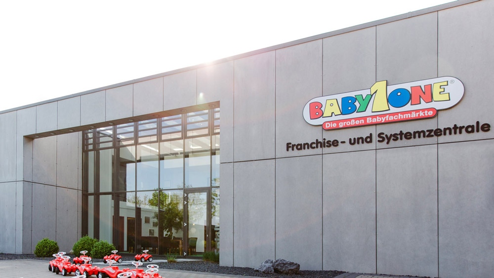 Modern building facade with the colorful "BabyOne" logo and the words "Franchise and System Headquarters." Several red toy cars are parked in front of the entrance.