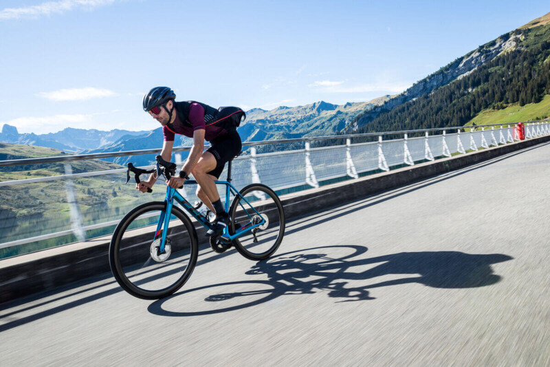 A cyclist in athletic clothing rides across a bridge with mountains in the background in sunny weather.