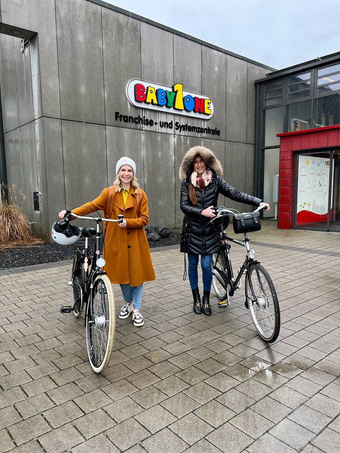 Two women are standing with bikes in front of the BabyOne franchise and system headquarters building.