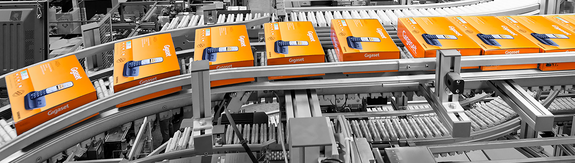 Several orange Gigaset packages travel on a conveyor belt through an industrial production facility. The surroundings are kept in shades of gray, which makes the colorful boxes stand out.