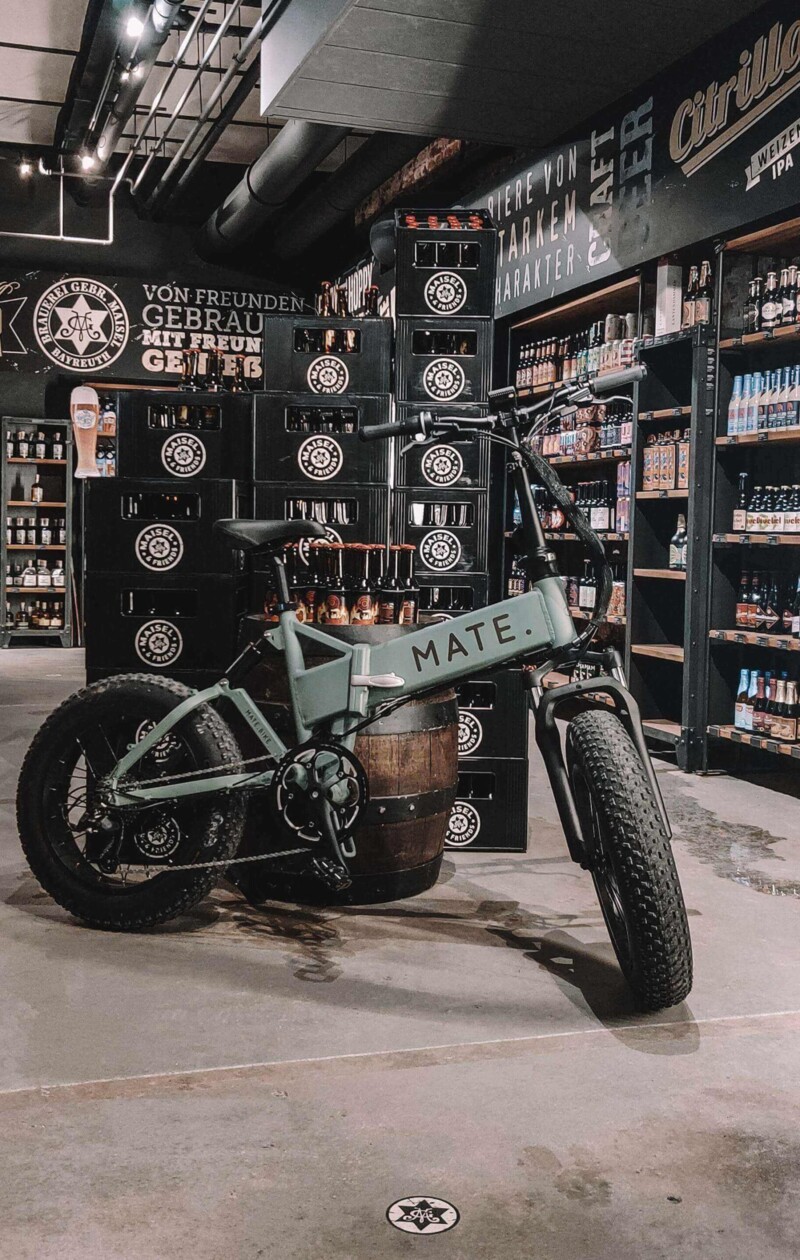 A gray MATE e-bike stands in front of a wooden barrel in a craft beer shop. Beer crates and shelves with various types of beer can be seen in the background.