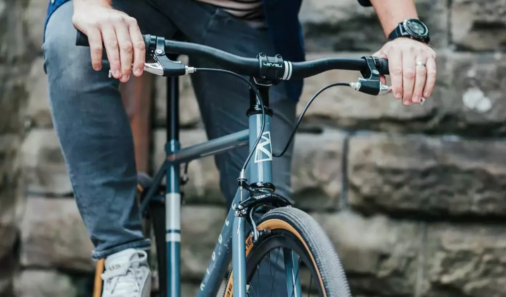 Close-up of a bicycle handlebar with the hands of a cyclist holding the bike. The rider is wearing gray pants, light-colored shoes, and a watch on his left wrist. Roughly hewn stone walls can be seen in the background.