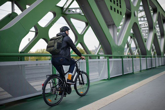 A man wearing a helmet and carrying a backpack rides a bike across a bridge with a green steel frame. The floor of the bridge is painted green, and the structure consists of diagonal and geometric elements.