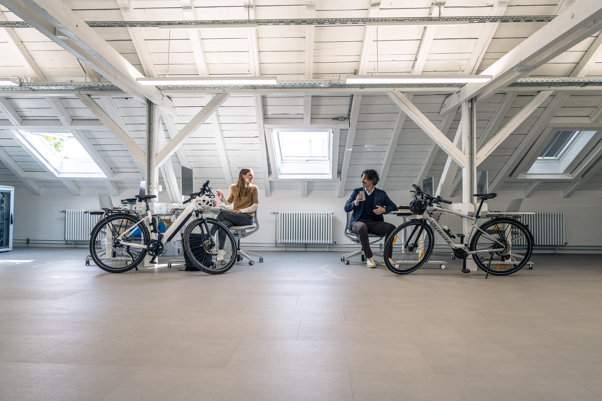 Two people are chatting in a bright attic office, each sitting next to an e-bike.
