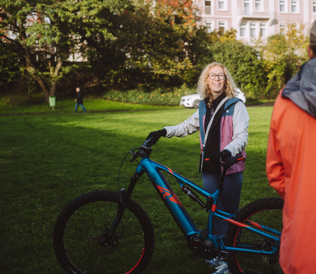 A woman with curly hair and glasses is standing on a green field next to a blue e-bike. Trees and buildings can be seen in the background. She is wearing a colorful jacket and smiling at a person on the right side of the picture.