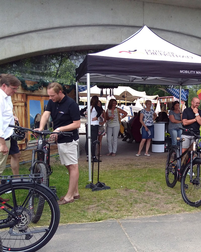 People are chatting and looking at bikes under a pavilion with the inscription "Deutsche Dienstrad" (German company bike). In the background, there are more people, a stand with decorations, and a large pavilion. The scene is set outdoors, possibly at an event or trade fair.