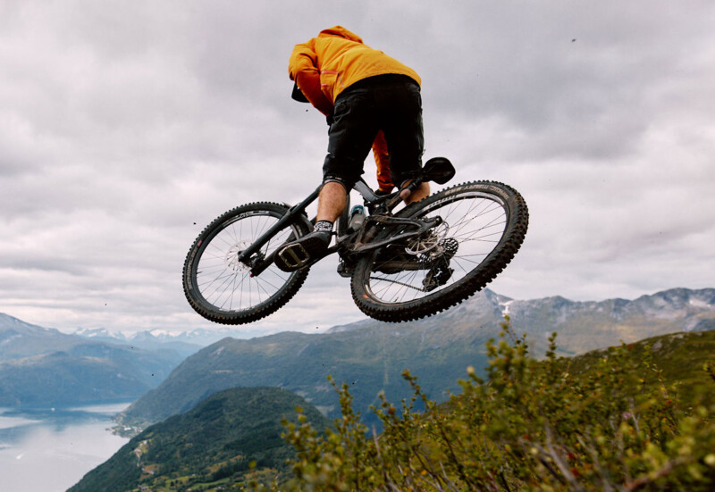 A mountain biker wearing an orange top jumps into the air with his bike, while a picturesque mountain landscape with a lake can be seen in the background.