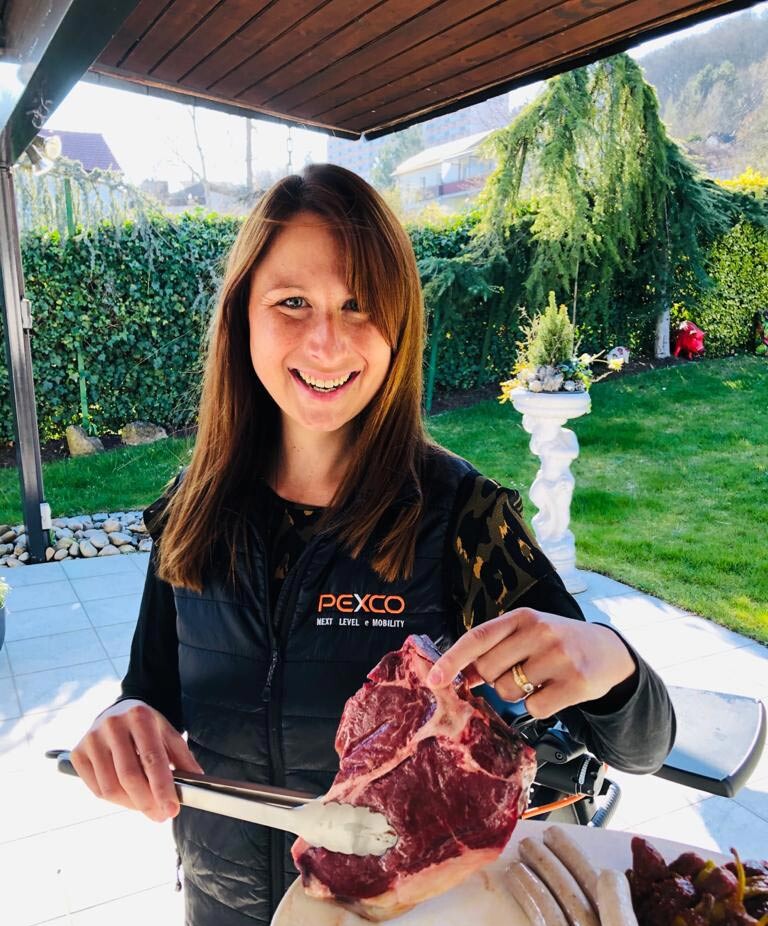 A woman is standing outside under a covered area, holding a large piece of raw meat with tongs. She smiles at the camera and is wearing a black vest with the words "Pexco - Next Level Mobility" printed on it. In the background, there is a well-kept garden with a lawn, bushes, and a decorative planter.