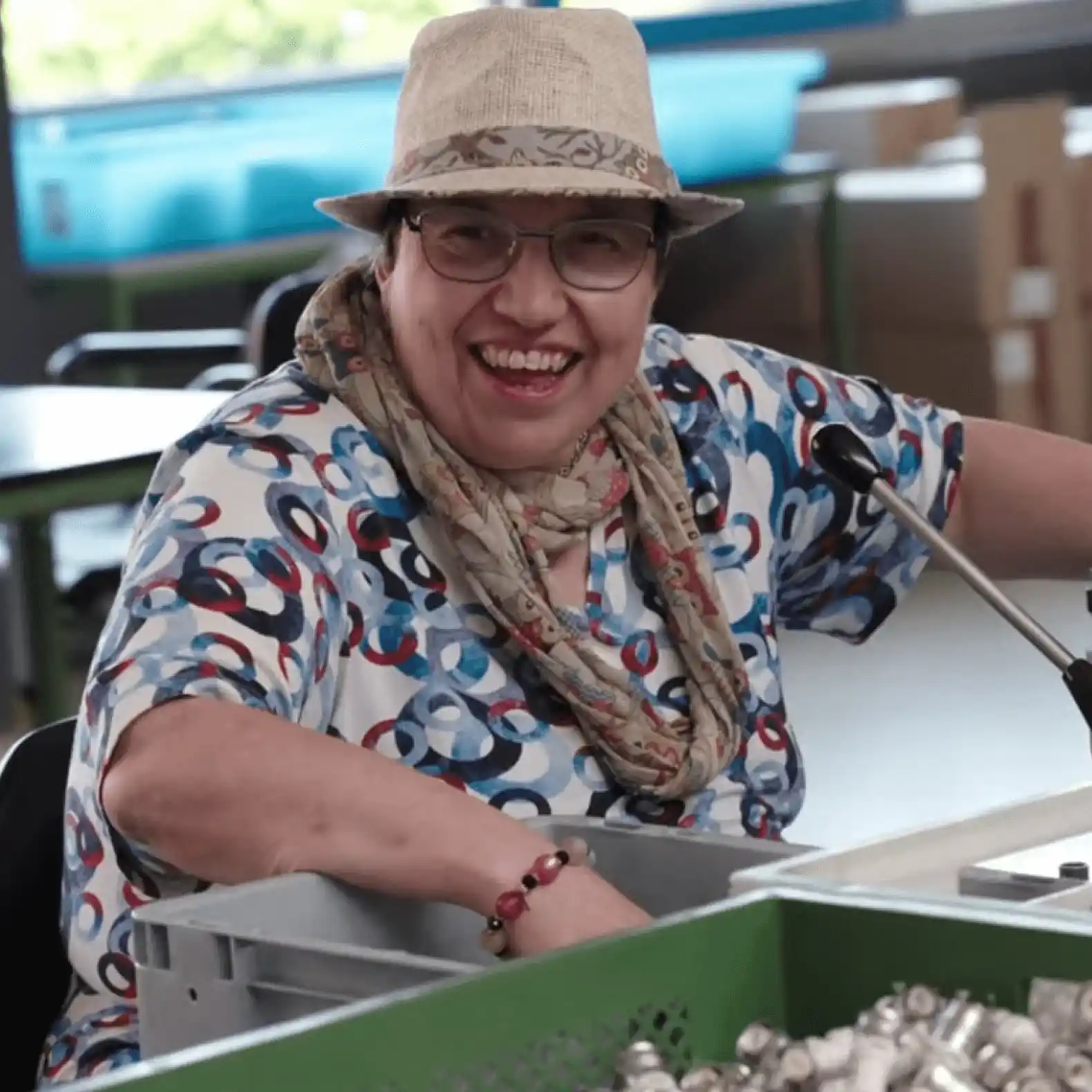 A smiling woman wearing a hat and glasses sits at a workstation sorting small items into a box. She is wearing a colorful top with a circular pattern and a scarf.