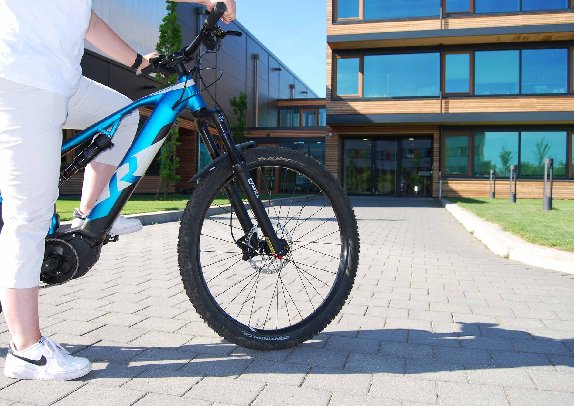 Person in white clothing on a blue bike, standing on a paved path in front of a modern building with a wooden facade and large windows.