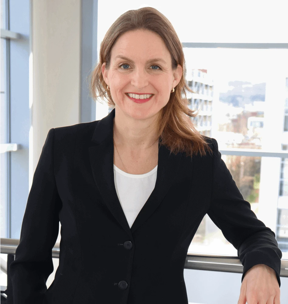 A smiling woman in a black blazer in front of a bright, modern office building in the background.