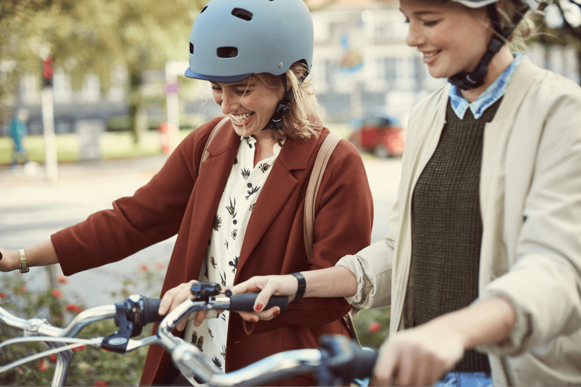 Two women wearing bicycle helmets smile as they stand side by side on their bikes.