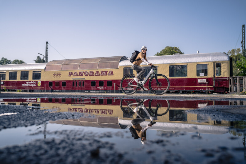 A cyclist rides past a historic panoramic train, its reflection visible in a puddle.