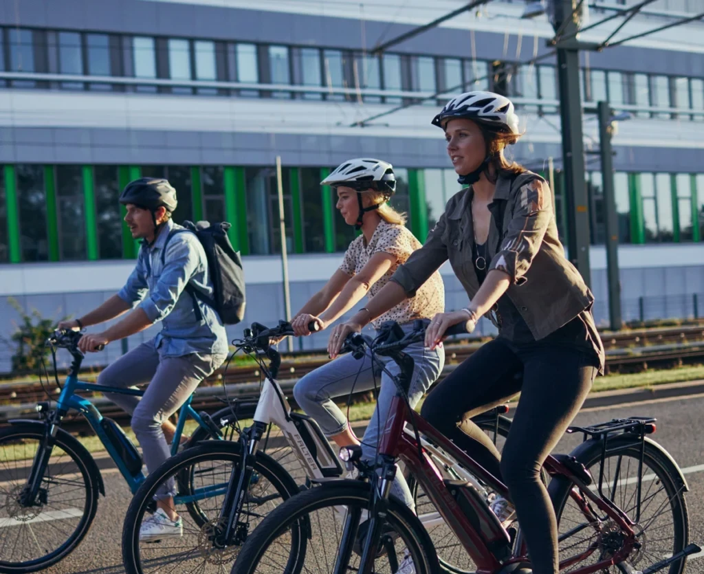 Three people wearing helmets ride e-bikes on a road in front of a modern building.