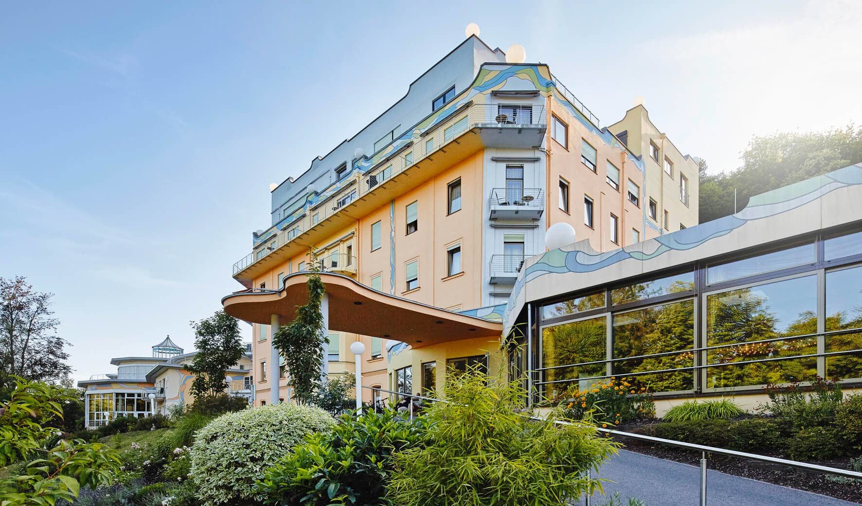 A multi-story, bright building with decorative wave patterns on the facade. In front of the building is a well-kept garden with lush plants. The sky is clear, and the scene feels calm and inviting.
