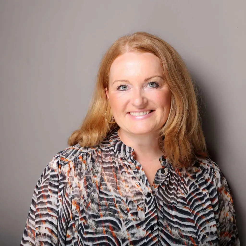 A smiling woman with shoulder-length, reddish-blonde hair stands in front of a gray background. She is wearing a patterned blouse in natural and orange tones.