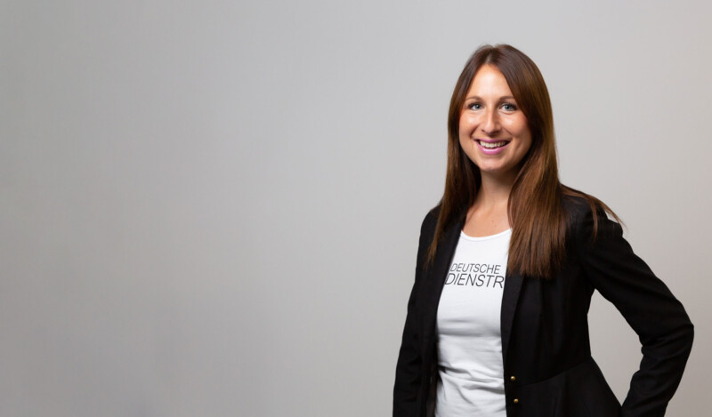A woman wearing a black blazer and a white T-shirt with "DEUTSCHE COMPANY BIKES" written on it, standing in front of a neutral gray background.