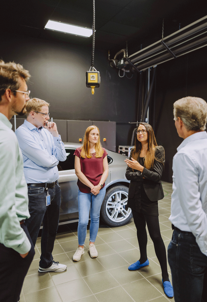 Fünf Personen stehen in einer Werkstatt um ein graues Auto und diskutieren. Eine Frau spricht gestikulierend, während die anderen zuhören.