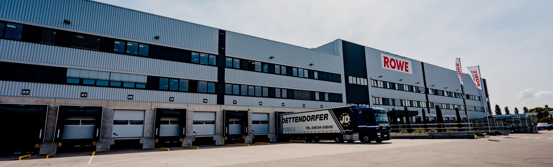 A large industrial building with the word "ROWE" written on the façade. In front of it, a truck belonging to the company "Dettendorfer" is parked at one of the loading ramps. Several roller doors for freight transport can be seen, and the sky is clear.