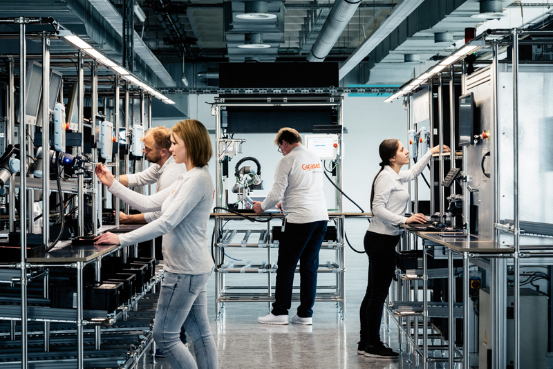 Several people working at workstations in a modern production area. The employees wear gray clothing while operating machines and computers to carry out production processes.